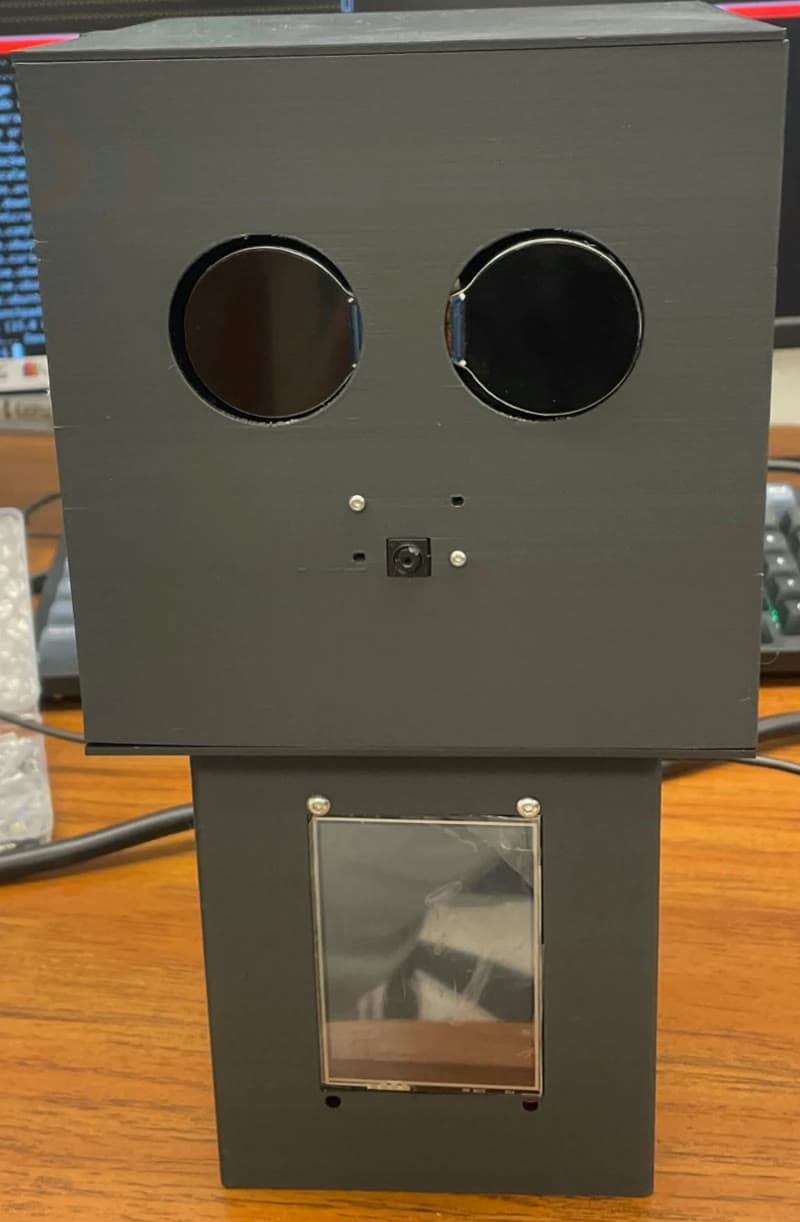The NanaBot prototype: a friendly white robot with round screen eyes and a touchscreen on its chest, greeting an older woman at home.